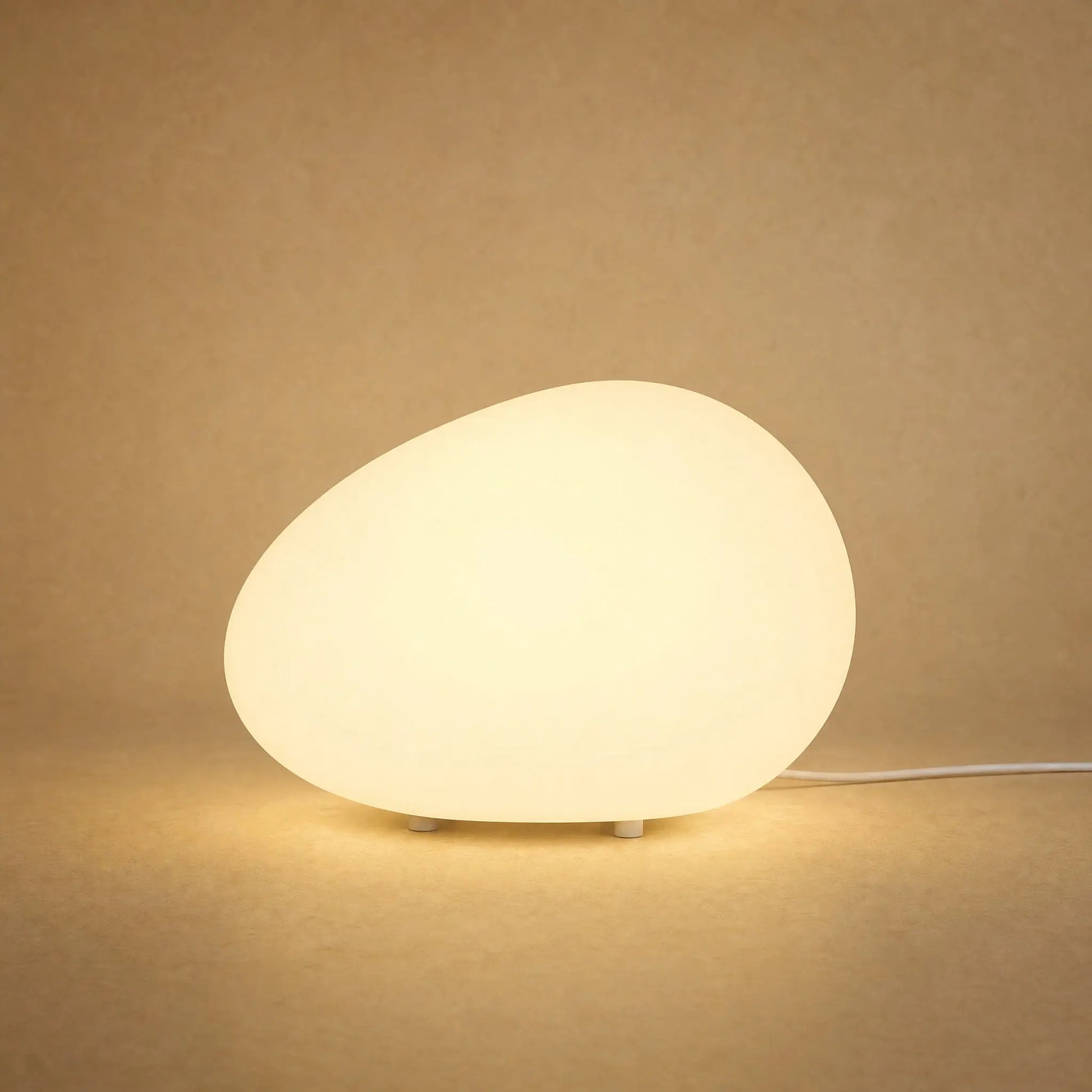Lampe à poser galet lumineux Sassolia allumée en blanc chaud sur fond minéral sable | Delvanie