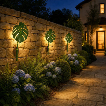 Trois appliques extérieures feuille de Monstera alignées et allumées le long d'un mur de jardin en pierre à la nuit tombante | Delvanie