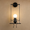 Applique murale noire Girotta, petit personnage assis sur sa balançoire, ampoule globe allumée | Delvanie
