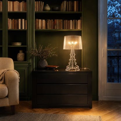 Lampe baroque Rivento transparente allumée sur une commode laquée noire devant une bibliothèque vert anglais | Delvanie